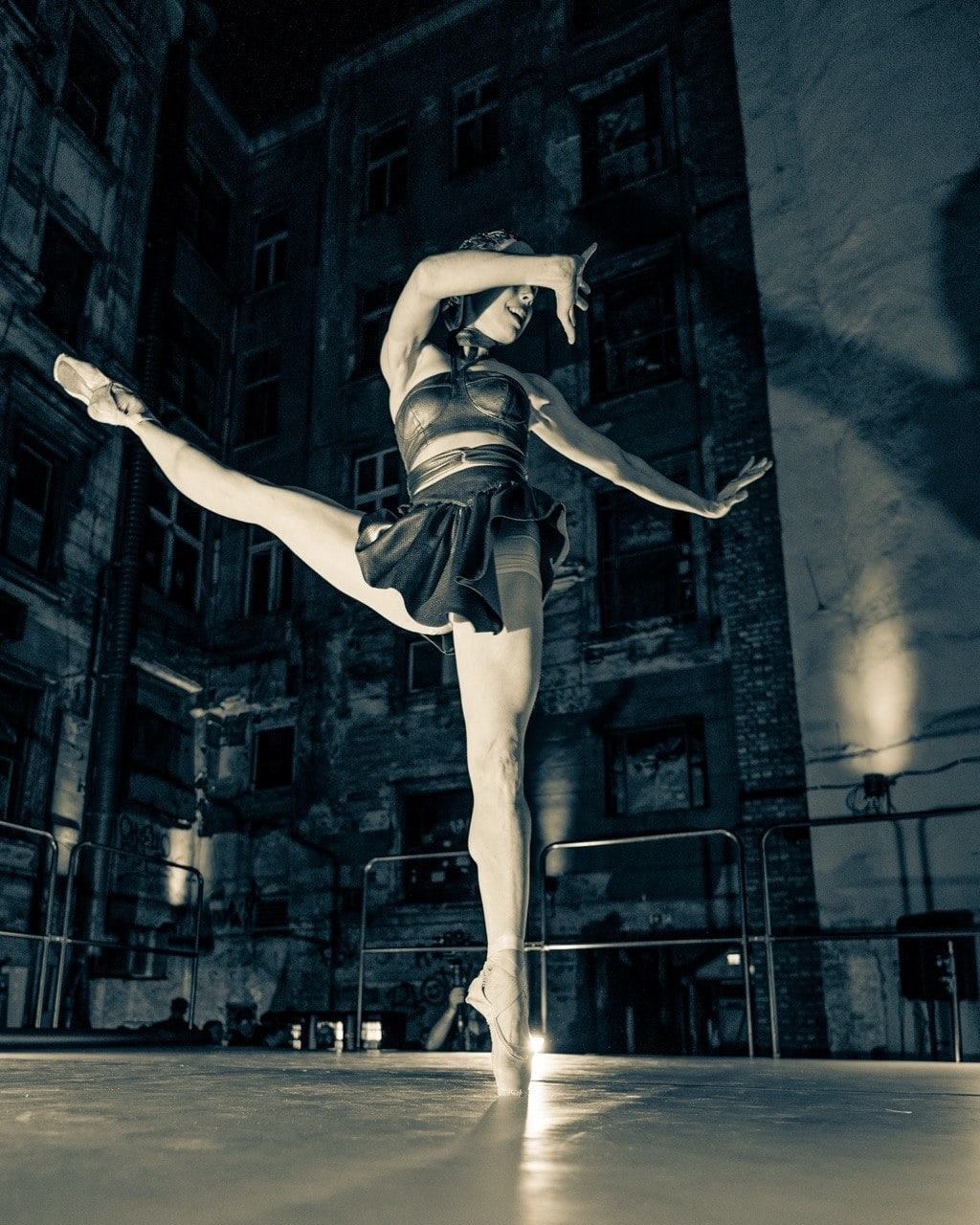 Ballet dancer in arabesque on the lit stage floor in the night courtyard, one arm shielding her face