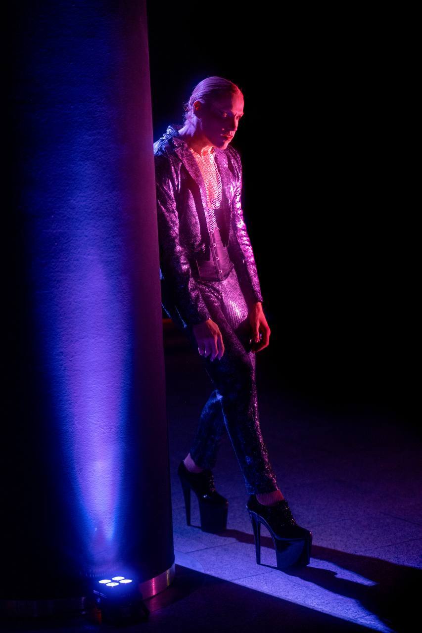 A performer in a sequined suit against a violet-lit column